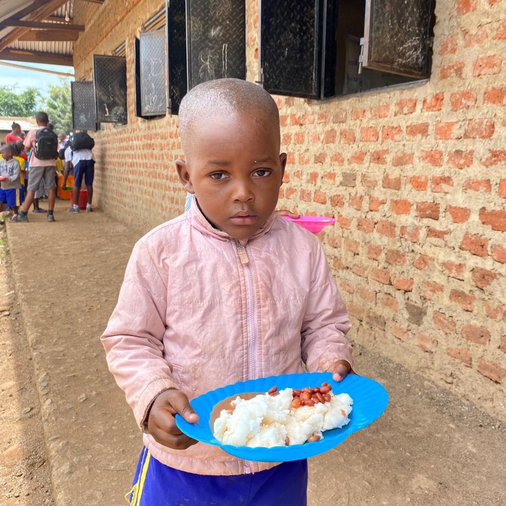 A young boy in a pink jacket holds a plate of rice at a school meal