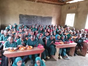 Pupils in green uniforms eat a meal at their desks in a crowded classroom